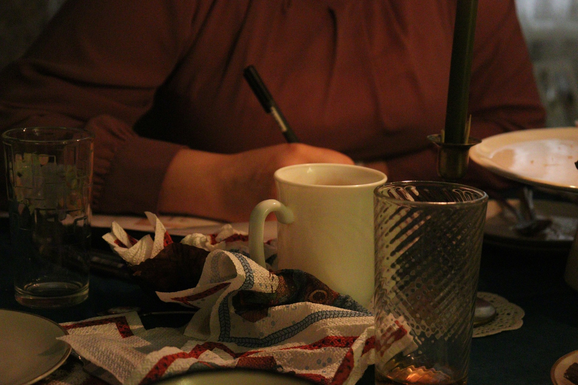 person writing at a dining table beside a mug and drinking glasses