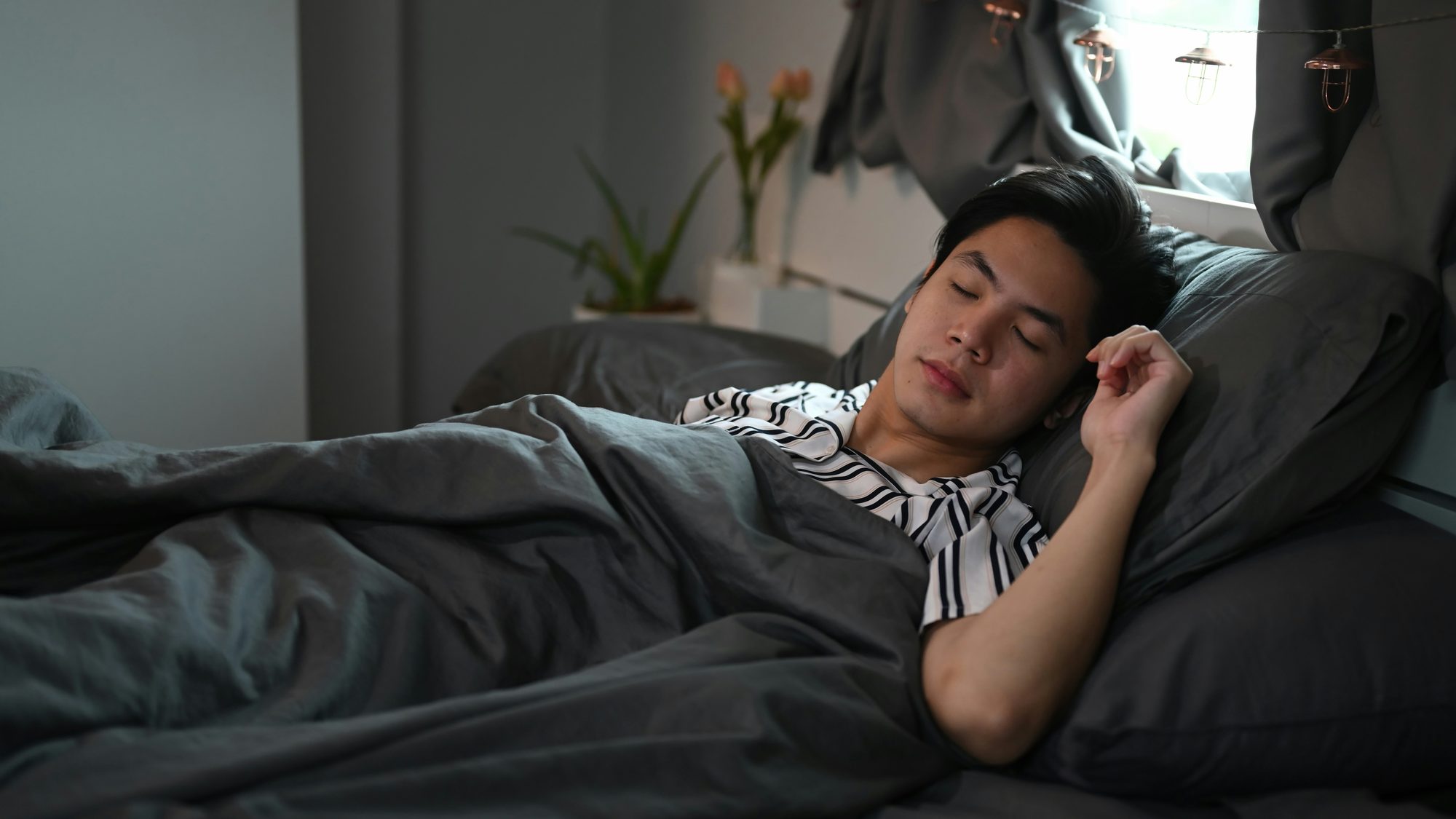 A young man asleep in bed under a grey duvet in a dim bedroom, with soft light coming through the curtains, illustrating the transition from Sunday night into Monday morning