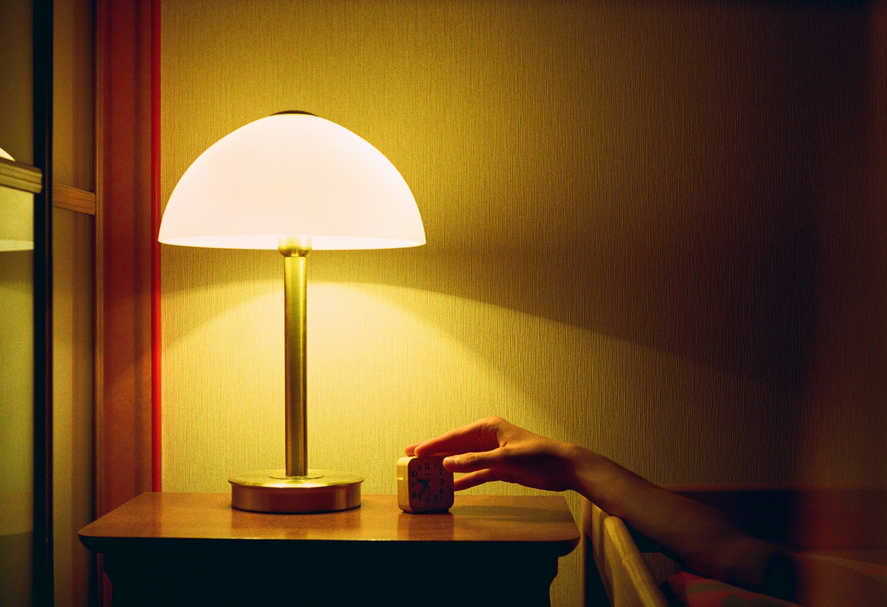A hand reaching toward a bedside alarm clock beside a warmly lit lamp