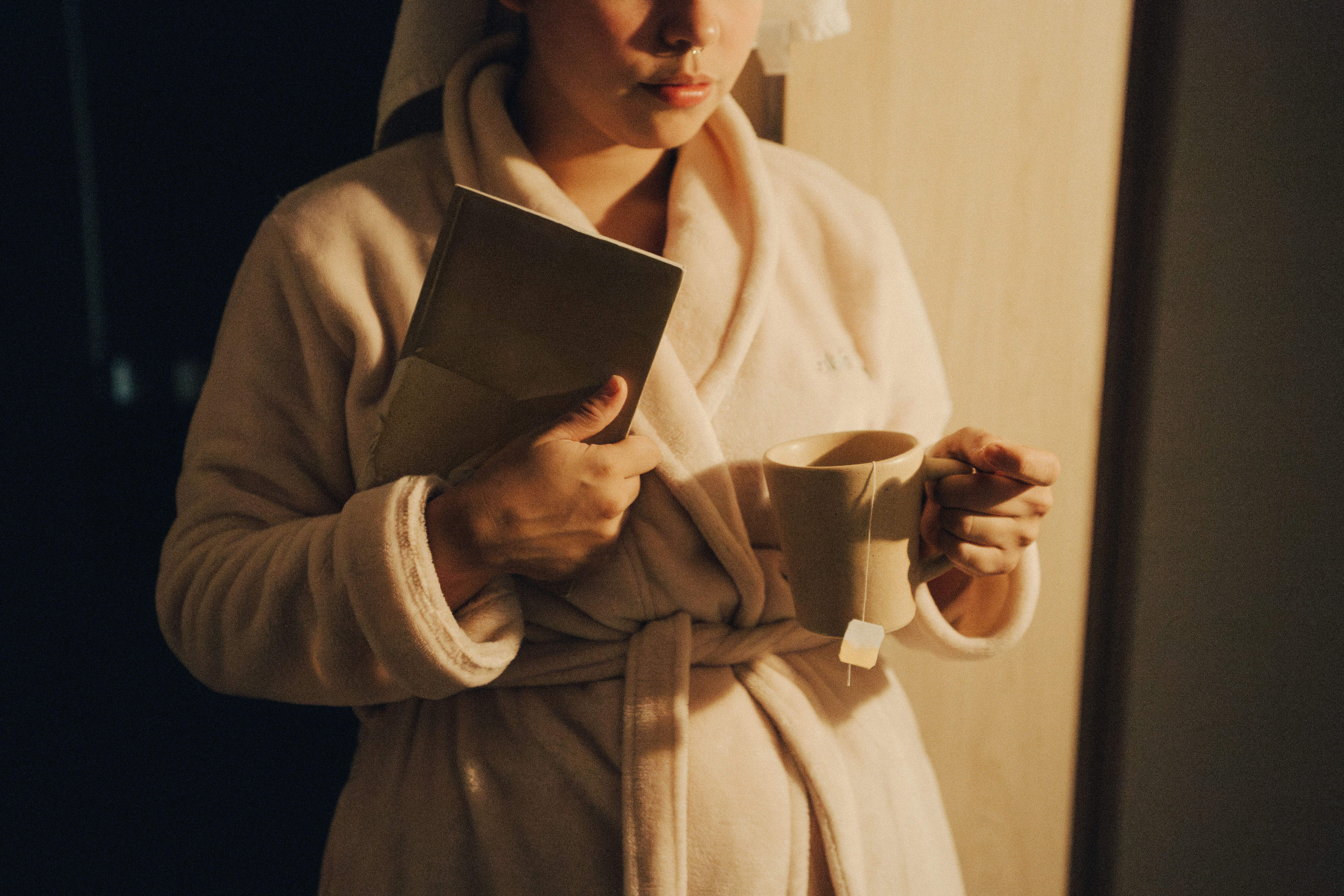 A person in a robe holding a book and a cup of tea in warm evening light, winding down as part of a bedtime routine